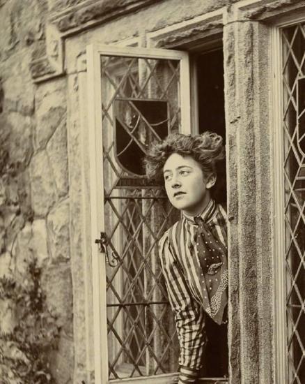 A black and white photograph showing a young woman leaning out of a window. 