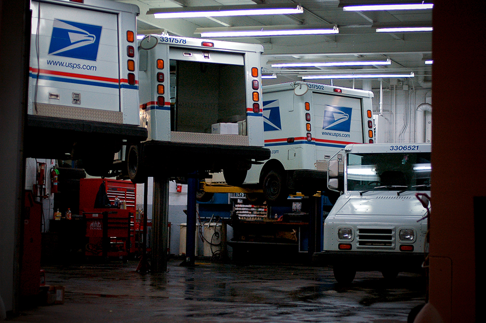 This is a photo of a car mechanic’s shop. There are three United States Postal Services trucks being serviced, and one not being serviced.